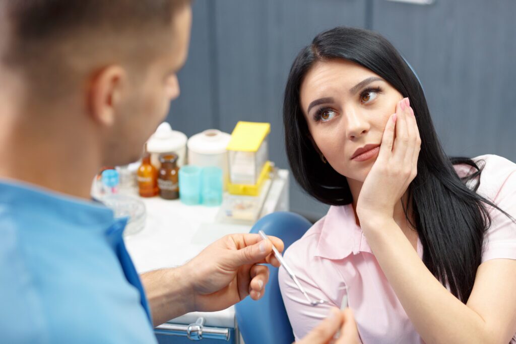 Female dental patient holding jaw in pain, seated in chair talking to dentist.