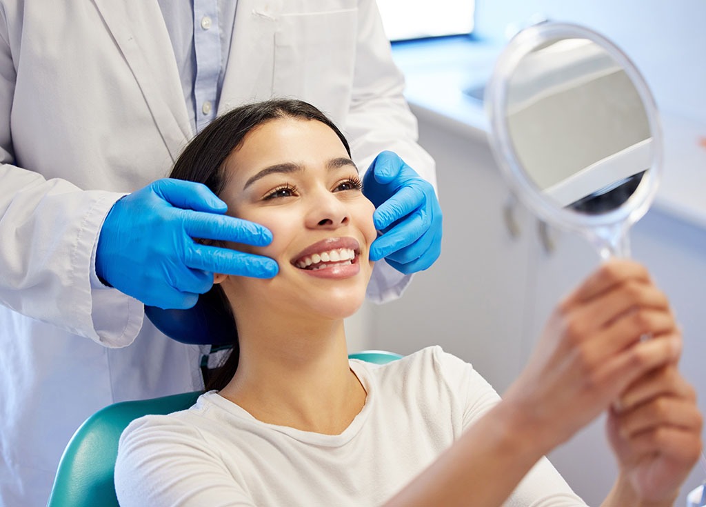 A woman in a dentist chair, holding a mirror up and smiling happily at the results of her teeth whitening.